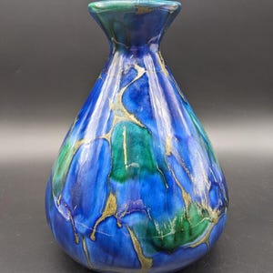 May include: A handcrafted ceramic vase with a unique design. The vase features a blend of blue and green hues, accented with gold-colored streaks. The vase has a wide base and a narrow neck, making it a decorative piece.