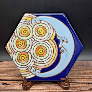 May include: A hexagonal ceramic tile featuring a stylized light blue crescent moon with a face, surrounded by concentric circles in shades of orange, yellow, and white against a deep blue background. The artist signature DYN is visible.