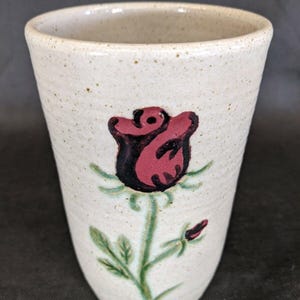 May include: A handmade ceramic tumbler with a cream-colored speckled finish. A large red rose with green leaves and stem is painted on the front. The tumbler has a slightly flared shape and a smooth rim.
