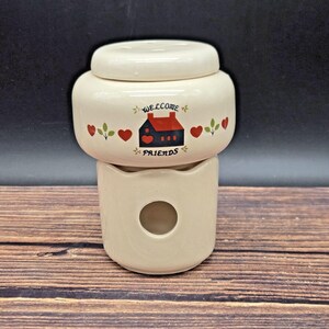 May include: Cream-colored ceramic jar with a lid, featuring a decorative design. The jar has a house illustration with the words "WELCOME FRIENDS" and red hearts. The jar sits on a matching base with a circular opening.