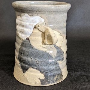 May include: Handmade ceramic vase with a unique design. The vase features a light gray exterior with textured, raised relief details in white, beige, and dark blue. The design appears to be a stylized figure. The vase has a cylindrical shape.