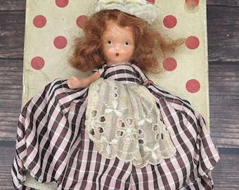 Puede incluir: Una muñeca vintage con pelo rojo, vestida con un vestido de cuadros rosa y marrón con una superposición de encaje blanco y un sombrero blanco. La muñeca está tumbada sobre un fondo blanco con lunares rojos.