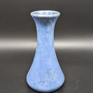 May include: A light blue ceramic vase with a textured surface. The vase has a flared top and a rounded base. The color is a soft, muted blue with subtle variations in tone, creating a unique, handcrafted appearance. The vase is suitable for displaying flowers.