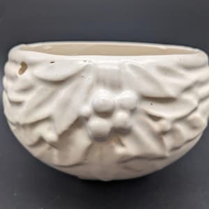 May include: White ceramic planter with a raised design of holly leaves and berries. The planter is a round shape and has a smooth, matte finish.