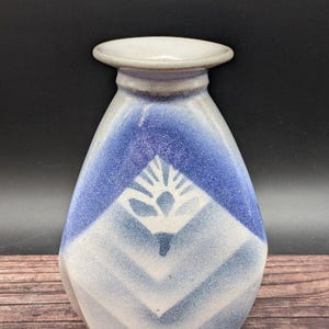 May include: A white ceramic vase with a blue and white geometric design. The vase has a rounded bottom and a narrow neck.