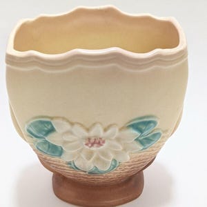 May include: A cream-colored ceramic vase with a scalloped rim and a floral design. The vase features a raised white flower with a pink center and teal leaves. The base is brown, and the overall design is vintage.