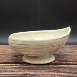 May include: Cream-colored ceramic serving dish with a boat-like shape, resting on a pedestal base. The dish features horizontal, textured, light brown stripes. The interior is smooth and the rim curves upwards.