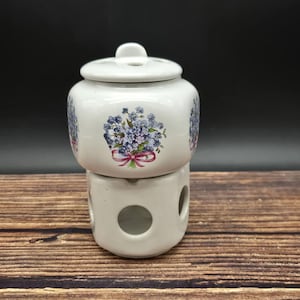 May include: A white ceramic jar with a lid, decorated with a blue floral design and a pink ribbon. The jar sits atop a matching base with circular openings. The background is dark, and the jar is on a wooden surface.