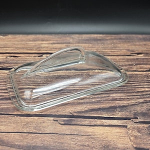 May include: A clear glass lid with a handle. The lid is rectangular with rounded corners and a raised handle on top. The glass is transparent, allowing a view of the surface beneath.
