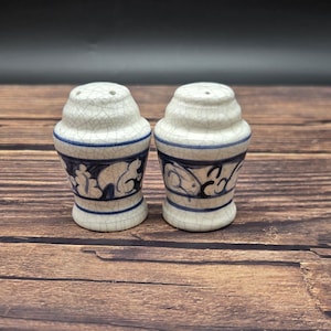 May include: A pair of ceramic salt and pepper shakers. The shakers are white with a crackle glaze and blue decorative bands. The lower portion of each shaker features a blue and white floral design.
