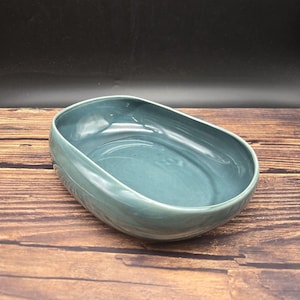 May include: A smooth, oval-shaped ceramic bowl with a glossy, muted teal-blue glaze. The bowl features a deep, rounded basin and a thick, curved rim, showcasing a consistent, solid color finish throughout the interior and exterior surfaces.