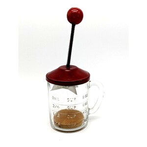 May include: A vintage red and glass hand-crank food mill with a glass measuring cup marked with 1/4 cup, 1/2 cup, 3/4 cup, and one cup. The mill has a red lid and a red knob on the top.