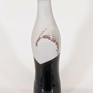May include: A ceramic bottle with a unique design. The top half is white with a textured, ribbed pattern and a maroon accent. The bottom half is glossy black. The bottle has a narrow neck and a rounded base.