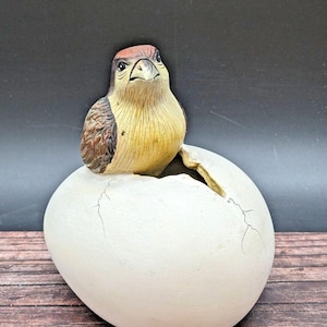 May include: A ceramic figurine of a bird hatching from a cracked egg. The bird is brown, yellow, and red with a detailed face. The egg is white with cracks and a rough texture.