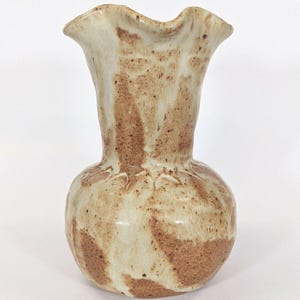 May include: A ceramic vase with a ruffled rim and a bulbous base. The vase has a speckled, earthy color palette of beige and brown, with darker brown markings. The vase is a decorative item.