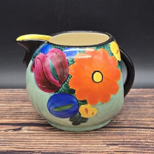 May include: A ceramic pitcher with a black handle and spout, adorned with vibrant floral designs. The pitcher features a light green base color, with large orange, pink, blue, and yellow flowers. The rim is beige, and the spout has a yellow accent.