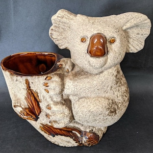 May include: A ceramic koala bear planter in shades of tan and brown. The koala is hugging a tree trunk-shaped planter with a brown interior. The koala has a brown nose and eyes. The planter has a textured finish.