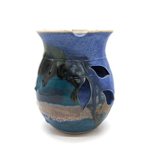 May include: A handcrafted ceramic oil warmer with a blue, teal, and brown color scheme. The vessel features leaf-shaped cutouts and a spout. The top rim is a lighter shade of blue.