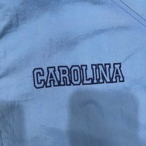 May include: Light blue fabric with the word "CAROLINA" embroidered in navy blue block letters. The fabric has a subtle sheen and a visible seam running diagonally across the top right corner.