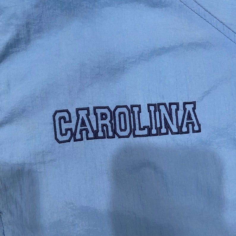 May include: Light blue fabric with the word "CAROLINA" embroidered in navy blue block letters. The fabric has a subtle sheen and a visible seam running diagonally across the top right corner.