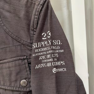 K&ouml;nnte beinhalten: Eine dunkelbraune Jacke aus strukturiertem Stoff mit einer Vordertasche und einem Metallknopf. Auf dem &Auml;rmel befindet sich ein wei&szlig;er Aufdruck mit dem Text 23 SUPPLY SQ. ROCKWELL FIELD AERONAUTIC SERVICE & REPAIR AME DIV. G75 CORONADO, CA ARMY AIR CORPS AVIREX.