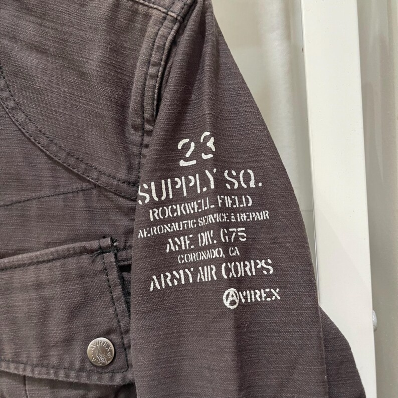 K&ouml;nnte beinhalten: Eine dunkelbraune Jacke aus strukturiertem Stoff mit einer Vordertasche und einem Metallknopf. Auf dem &Auml;rmel befindet sich ein wei&szlig;er Aufdruck mit dem Text 23 SUPPLY SQ. ROCKWELL FIELD AERONAUTIC SERVICE & REPAIR AME DIV. G75 CORONADO, CA ARMY AIR CORPS AVIREX.