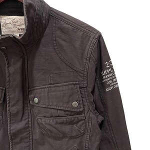 K&ouml;nnte beinhalten: Eine dunkelbraune Utility-Jacke mit hohem Kragen, zwei Brusttaschen mit Druckknopfverschluss und einem wei&szlig;en Grafikdruck auf dem &Auml;rmel. Das Etikett am Innenkragen tr&auml;gt die Aufschrift Armed Forces Manufacturer Avirex mit Text.