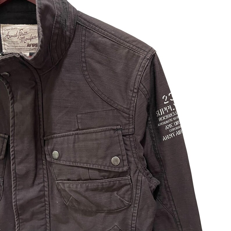 K&ouml;nnte beinhalten: Eine dunkelbraune Utility-Jacke mit hohem Kragen, zwei Brusttaschen mit Druckknopfverschluss und einem wei&szlig;en Grafikdruck auf dem &Auml;rmel. Das Etikett am Innenkragen tr&auml;gt die Aufschrift Armed Forces Manufacturer Avirex mit Text.