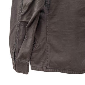 K&ouml;nnte beinhalten: Eine dunkel anthrazitfarbene Langarmjacke mit strukturiertem Kragen, vertikalen Nahtdetails und einem &Auml;rmelb&uuml;ndchen, das mit zwei metallischen Druckkn&ouml;pfen befestigt ist. Das Kleidungsst&uuml;ck besteht aus einem strukturierten Stoff mit sichtbaren N&auml;hten.