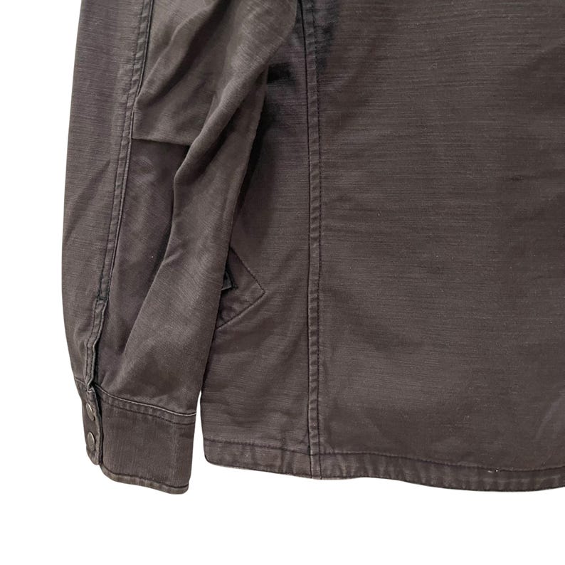 K&ouml;nnte beinhalten: Eine dunkel anthrazitfarbene Langarmjacke mit strukturiertem Kragen, vertikalen Nahtdetails und einem &Auml;rmelb&uuml;ndchen, das mit zwei metallischen Druckkn&ouml;pfen befestigt ist. Das Kleidungsst&uuml;ck besteht aus einem strukturierten Stoff mit sichtbaren N&auml;hten.