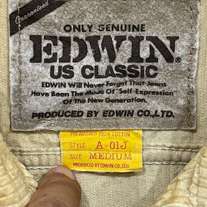 May include: A close-up shot of a beige fabric label with the text "EDWIN US CLASSIC" and "PREWASHED 100% COTTON". The label also shows the style "A-01J" and size "MEDIUM". The label is attached to a beige corduroy fabric.
