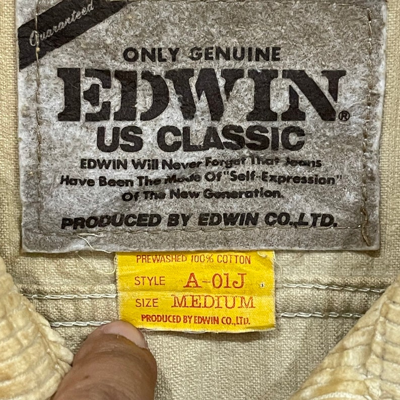 May include: A close-up shot of a beige fabric label with the text "EDWIN US CLASSIC" and "PREWASHED 100% COTTON". The label also shows the style "A-01J" and size "MEDIUM". The label is attached to a beige corduroy fabric.