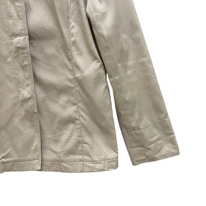 May include: Close-up of a beige jacket. The jacket has a tailored design with visible seams and a button closure. The sleeve is visible, showing a cuff detail. The fabric has a subtle sheen, suggesting a satin or similar material.