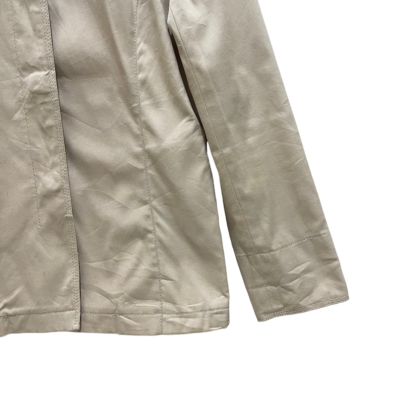 May include: Close-up of a beige jacket. The jacket has a tailored design with visible seams and a button closure. The sleeve is visible, showing a cuff detail. The fabric has a subtle sheen, suggesting a satin or similar material.