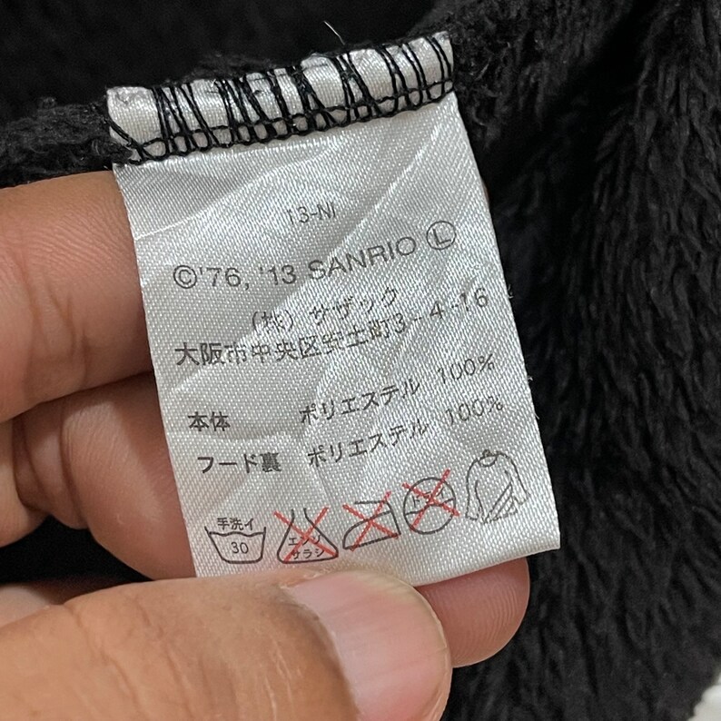 Peut inclure: Une &eacute;tiquette blanche avec du texte noir et des symboles. L'&eacute;tiquette indique "&copy;'76, '13 SANRIO (L)" et "大阪市中央区安土町3-4-16". L'&eacute;tiquette indique &eacute;galement "本体 ポリエステル 100%" et "フード裏 ポリエステル 100%". Il y a des symboles pour le lavage &agrave; la main &agrave; 30 degr&eacute;s Celsius et pour ne pas utiliser d'eau de Javel ou de s&egrave;che-linge.