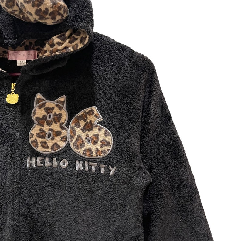 Peut inclure: Sweat-shirt &agrave; capuche en polaire noir avec une doublure de capuche en imprim&eacute; l&eacute;opard. Le devant pr&eacute;sente un grand motif brod&eacute; des chiffres "86" en imprim&eacute; l&eacute;opard avec le texte "HELLO KITTY" en dessous.