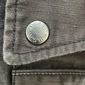 K&ouml;nnte beinhalten: Ein dunkelgraues Kleidungsst&uuml;ck aus Stoff mit einem runden, silberfarbenen Metallknopf. Der Knopf ist mit dem Text AVIREX U.S.A. und einem kleinen kreisf&ouml;rmigen Logo gepr&auml;gt. Der Stoff hat eine sichtbare Webstruktur mit dunklen N&auml;hten.