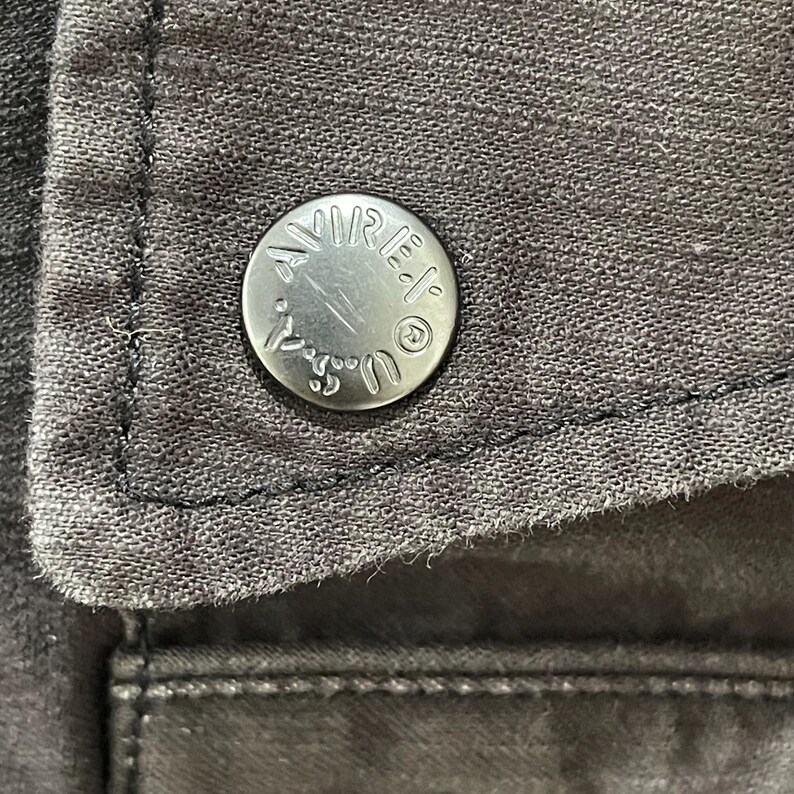 K&ouml;nnte beinhalten: Ein dunkelgraues Kleidungsst&uuml;ck aus Stoff mit einem runden, silberfarbenen Metallknopf. Der Knopf ist mit dem Text AVIREX U.S.A. und einem kleinen kreisf&ouml;rmigen Logo gepr&auml;gt. Der Stoff hat eine sichtbare Webstruktur mit dunklen N&auml;hten.