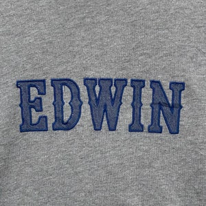 May include: Close-up of a gray fabric with the embroidered word "EDWIN" in blue. The letters are in a bold, stylized font, and the stitching is visible. The fabric has a textured appearance.