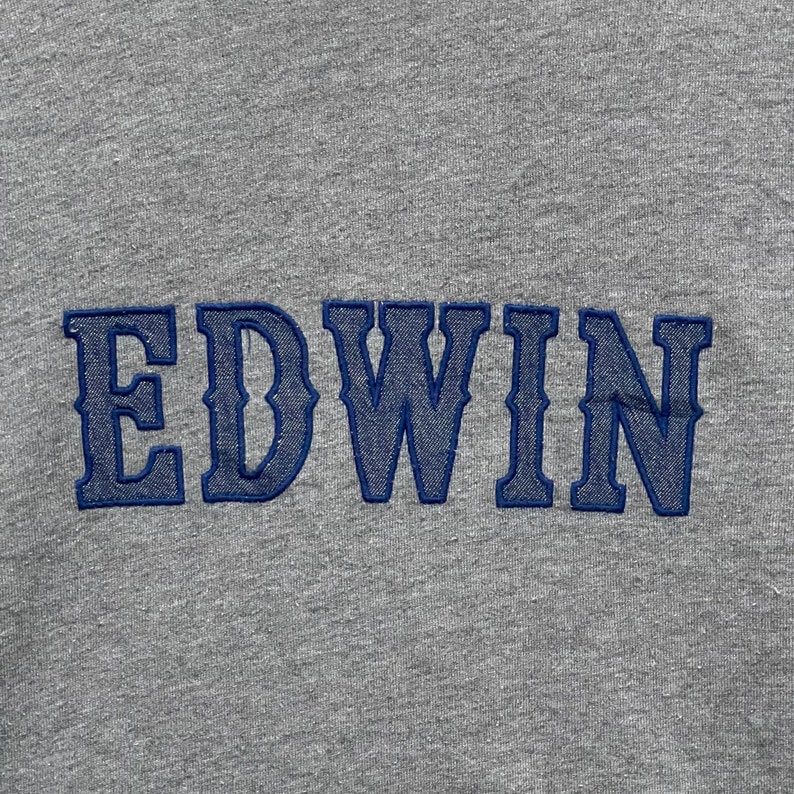 May include: Close-up of a gray fabric with the embroidered word "EDWIN" in blue. The letters are in a bold, stylized font, and the stitching is visible. The fabric has a textured appearance.
