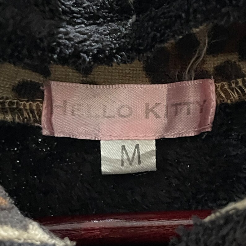 Peut inclure: Une &eacute;tiquette rose Hello Kitty avec le texte "HELLO KITTY" en lettres noires. Une &eacute;tiquette blanche avec la lettre "M" en lettres noires est attach&eacute;e &agrave; l'&eacute;tiquette rose.