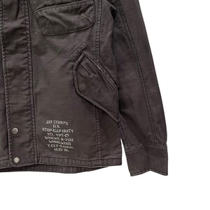 K&ouml;nnte beinhalten: Eine dunkelbraune Jacke mit einer Fronttasche mit Klappe, einer Brusttasche und Metalldruckkn&ouml;pfen. Auf der unteren Vorderseite befindet sich wei&szlig; gedruckter Text: AIR CORPS U.S. STOP-GAP DUTY STL. YPT-15 WRIGHT R-760 WHIRLWIND 7-CLY RADIAL H39 SL.