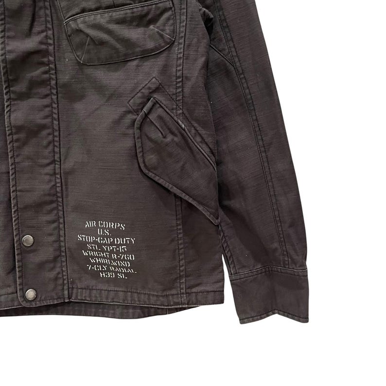 K&ouml;nnte beinhalten: Eine dunkelbraune Jacke mit einer Fronttasche mit Klappe, einer Brusttasche und Metalldruckkn&ouml;pfen. Auf der unteren Vorderseite befindet sich wei&szlig; gedruckter Text: AIR CORPS U.S. STOP-GAP DUTY STL. YPT-15 WRIGHT R-760 WHIRLWIND 7-CLY RADIAL H39 SL.