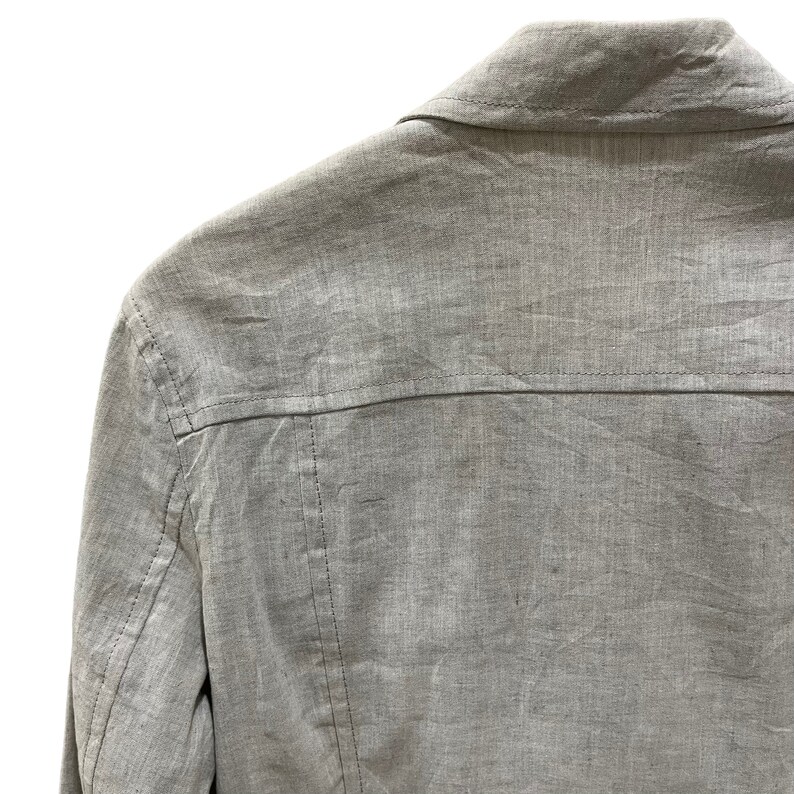 Puede incluir: Primer plano de una chaqueta gris claro, mostrando el cuello y la parte trasera. La tela tiene un aspecto texturizado con costuras visibles a lo largo de las costuras. La chaqueta parece estar hecha de un material ligero.