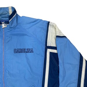 May include: Light blue Carolina jacket with white and navy blue accents. The jacket features the word "CAROLINA" in navy blue lettering. A Starter logo is visible on the upper left chest.