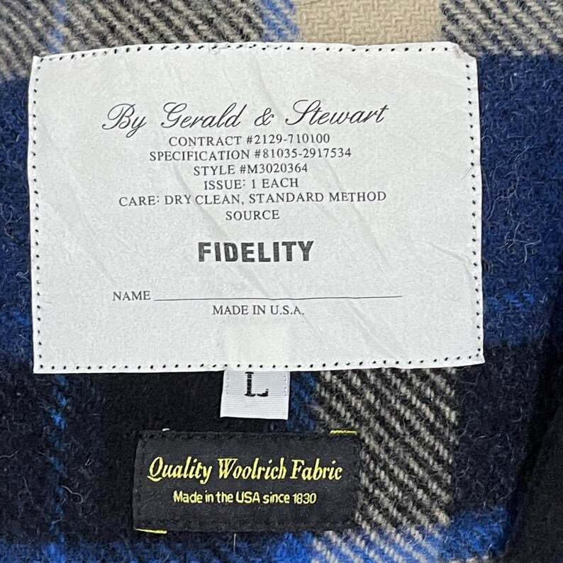 May include: A white rectangular garment label with black text reading By Gerald & Stewart, Contract #2129-710100, Specification #81035-2917534, Style #M3020364, Issue: 1 Each, Care: Dry Clean, Standard Method, Fidelity, Name, and Made in U.S.A.
