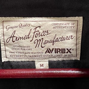 K&ouml;nnte beinhalten: Ein rechteckiges, cremefarbenes Stoffetikett mit braunem Text, das auf ein schwarzes Kleidungsst&uuml;ck gen&auml;ht ist. Das Etikett tr&auml;gt die Aufschrift Genuine Quality, Armed Forces Manufacturer, Certificate of Authenticity, Guarantee Good For One, Avirex und Established in 1975.