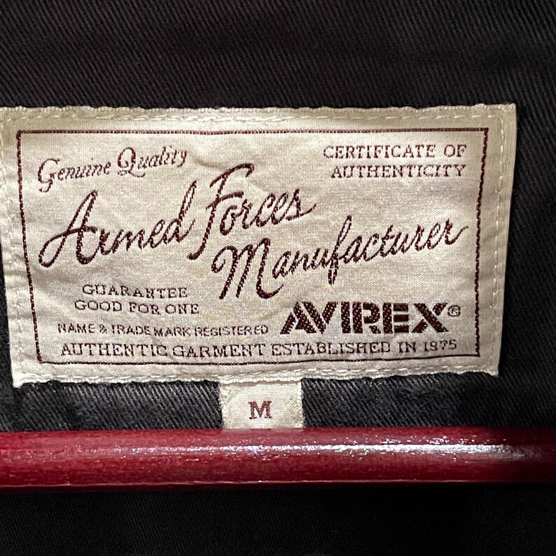 K&ouml;nnte beinhalten: Ein rechteckiges, cremefarbenes Stoffetikett mit braunem Text, das auf ein schwarzes Kleidungsst&uuml;ck gen&auml;ht ist. Das Etikett tr&auml;gt die Aufschrift Genuine Quality, Armed Forces Manufacturer, Certificate of Authenticity, Guarantee Good For One, Avirex und Established in 1975.