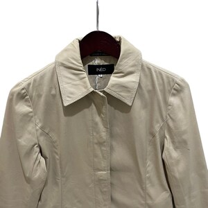 May include: A tan-colored IN&Egrave;D jacket hangs on a wooden hanger. The jacket has a collared neckline and a button-down front. The brand name, IN&Egrave;D, is displayed on a black tag at the collar. The jacket is made of a smooth, lightweight material.