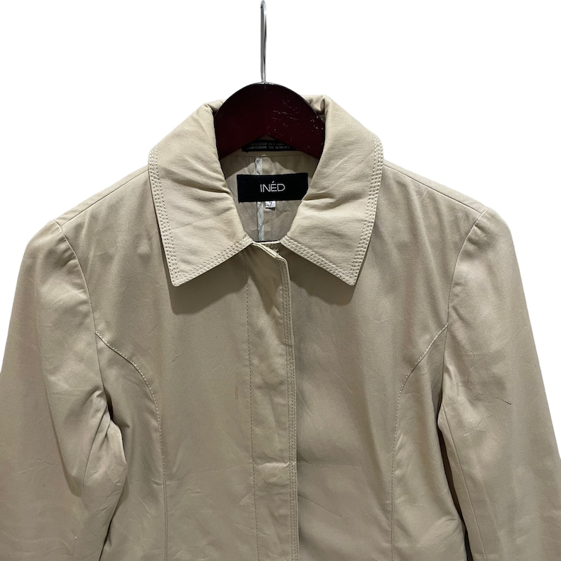 May include: A tan-colored IN&Egrave;D jacket hangs on a wooden hanger. The jacket has a collared neckline and a button-down front. The brand name, IN&Egrave;D, is displayed on a black tag at the collar. The jacket is made of a smooth, lightweight material.